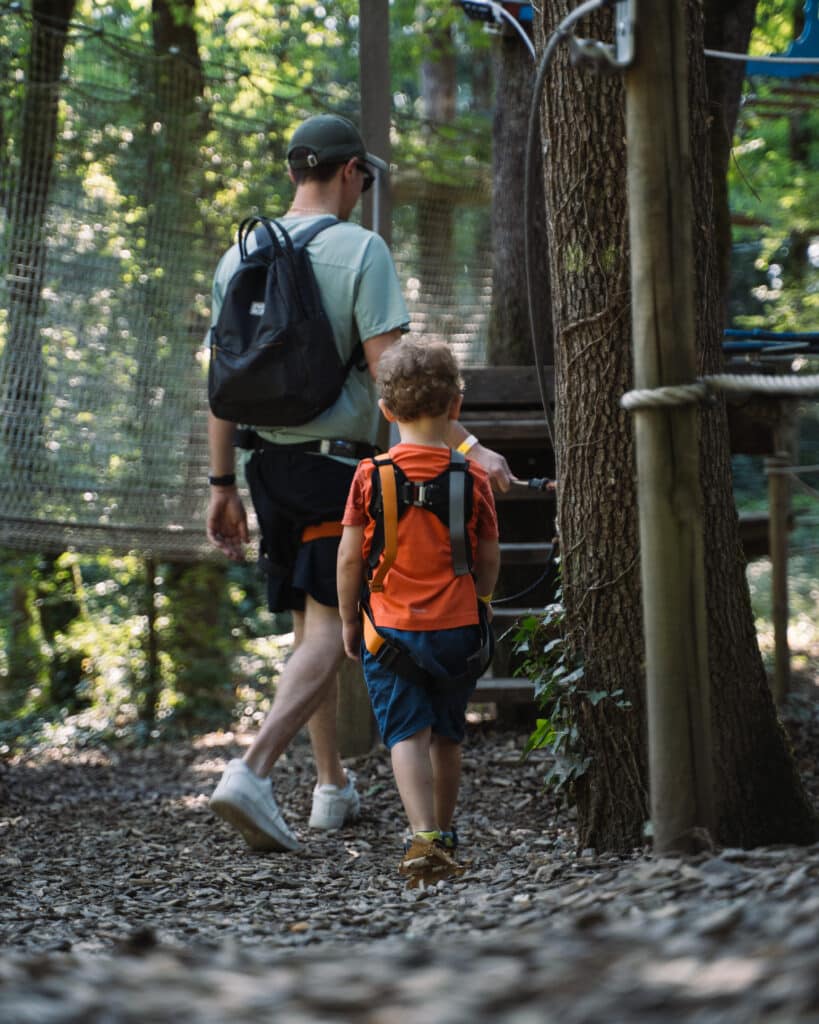 Accrobranche en famille : aventure et escalade en forêt Homme et enfant avec harnais marchant vers un parcours d'accrobranche en forêt sous le soleil.
