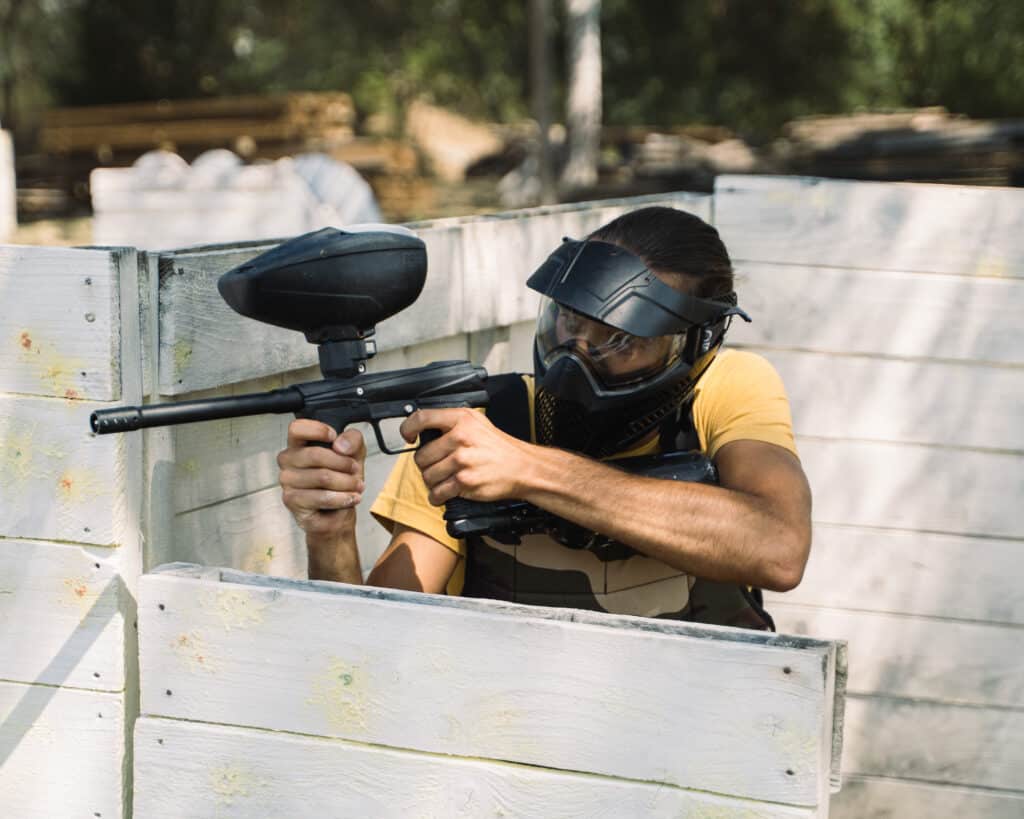 Paintball : Joueur en action derrière une palissade Joueur de paintball masqué visant avec son marqueur noir derrière un abri en bois blanc.