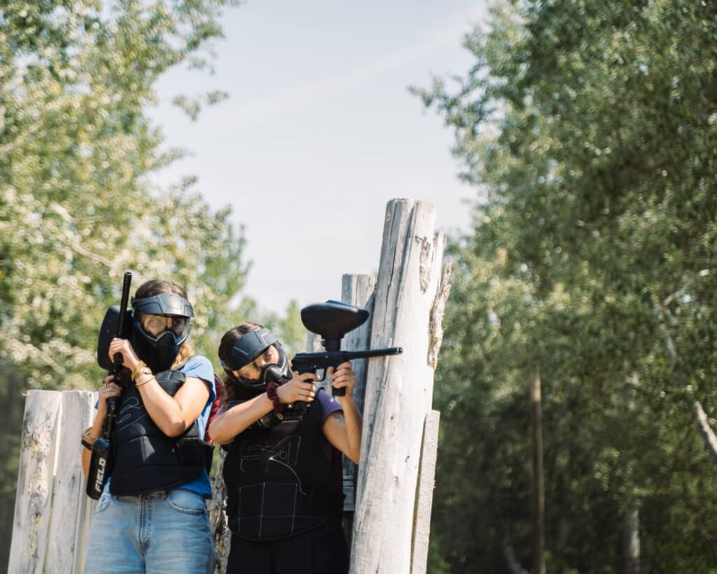 Paintball en Forêt : Action et Adrénaline entre Amies Deux femmes masquées en tenue de protection visent avec des lanceurs de paintball derrière une palissade.