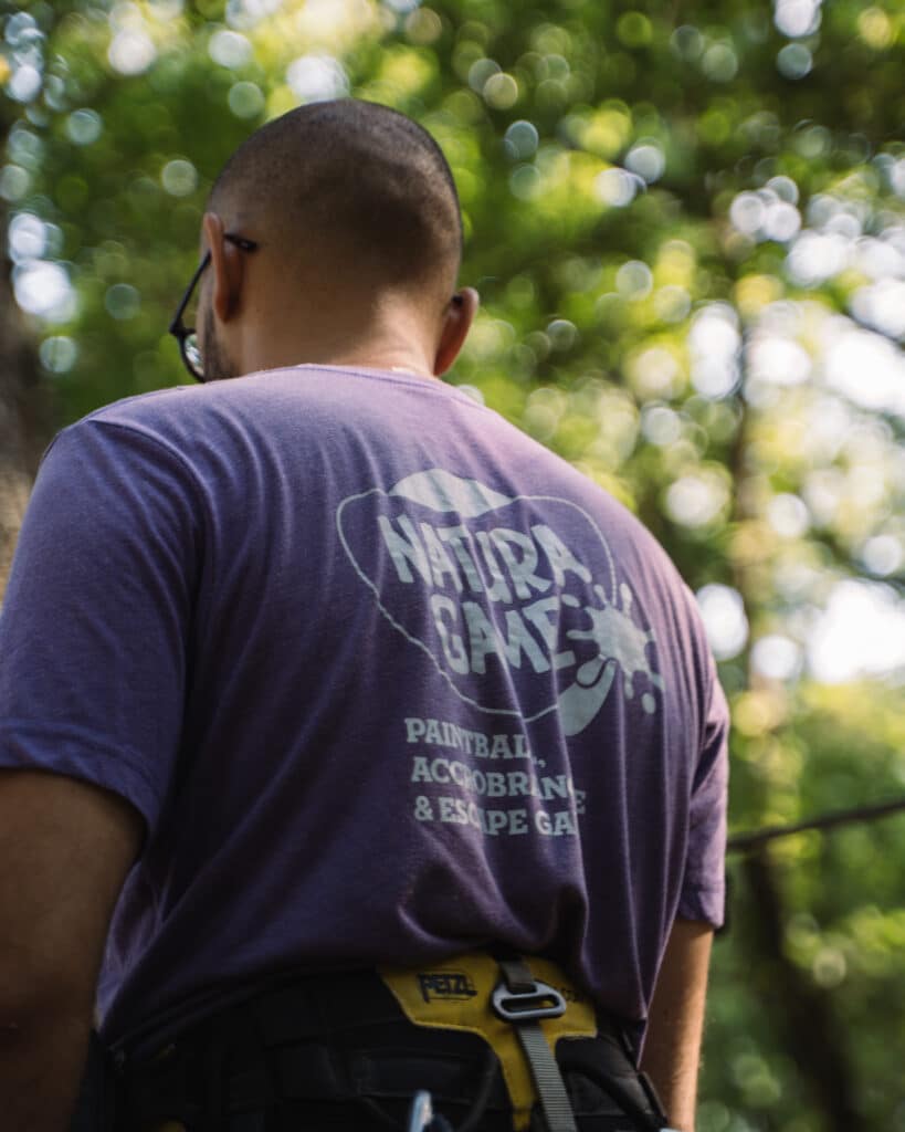 Natura Game : Paintball et Accrobranche en pleine nature Homme de dos, t-shirt Natura Game Paintball Accrobranche et baudrier Petzl, prêt pour l'aventure en forêt.